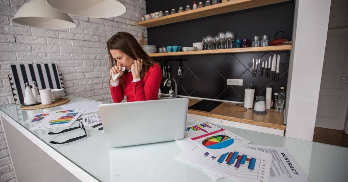 A white woman sits at a counter in front of a computer while sneezing